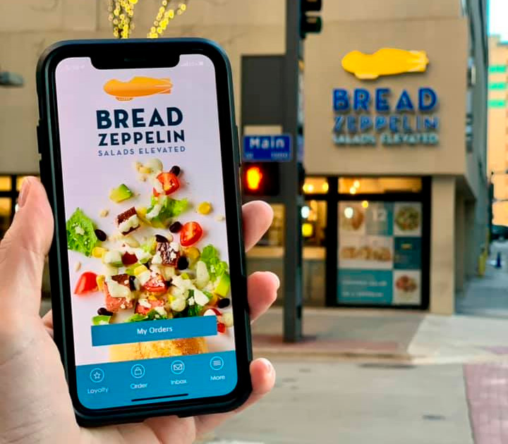 Man Holding Smartphone with Bread Zeppelin App: A man smiling while holding a smartphone displaying the Bread Zeppelin app, showcasing user-friendly features and menu options for ordering fresh salads and sandwiches.